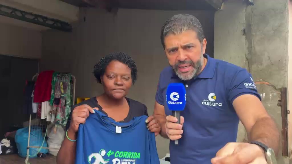 Tia Lu segura uma camisa azul da 4ª Corrida Rex ao lado do repórter da Rádio Cultura