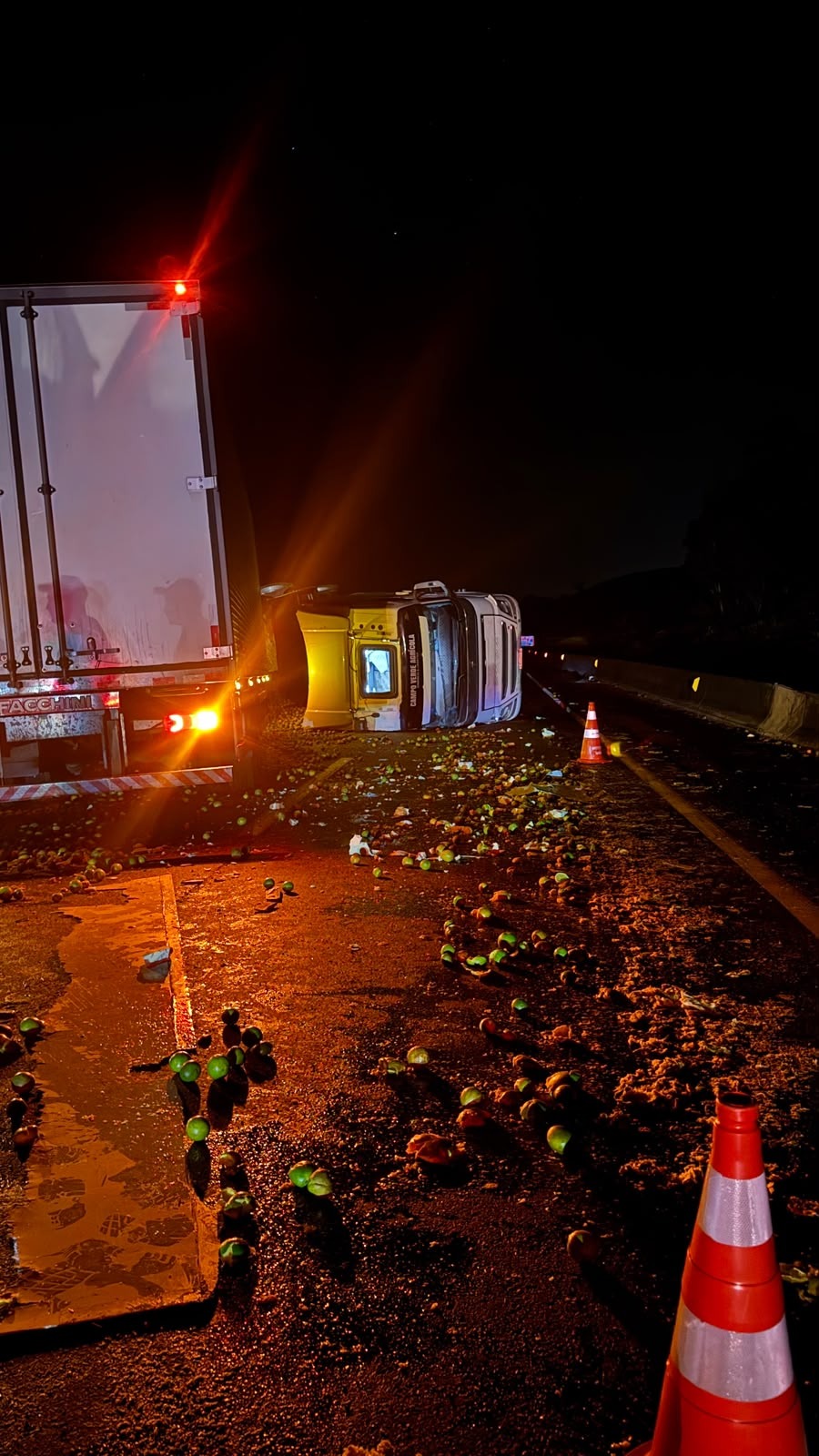 Carreta tombada de lado na pista da BR-381, com laranjas espalhadas pelo asfalto e cones de sinalização