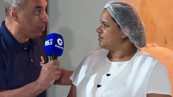 Repórter da Rádio Cultura com microfone entrevista mulher de touca e avental branco na cozinha da festa