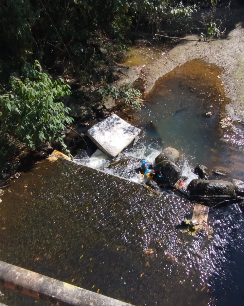 Contêiner de lixo tomado por sacolas e resíduos que se espalham pela calçada, perto do Ribeirão da Água Limpa
